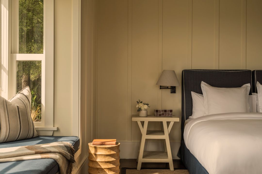 A hotel suite with neutral interiors, plush bed and window seat with sun streaming in.